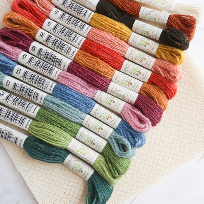 DMC Eco Vita organic wool thread skeins arranged diagonally by color on a textured cream aida fabric.